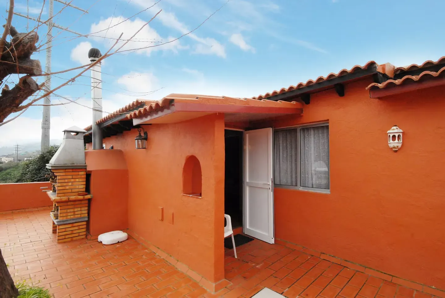 Una casita de color terracota con una barbacoa de ladrillo, una puerta blanca y un patio con baldosas bajo un cielo azul.