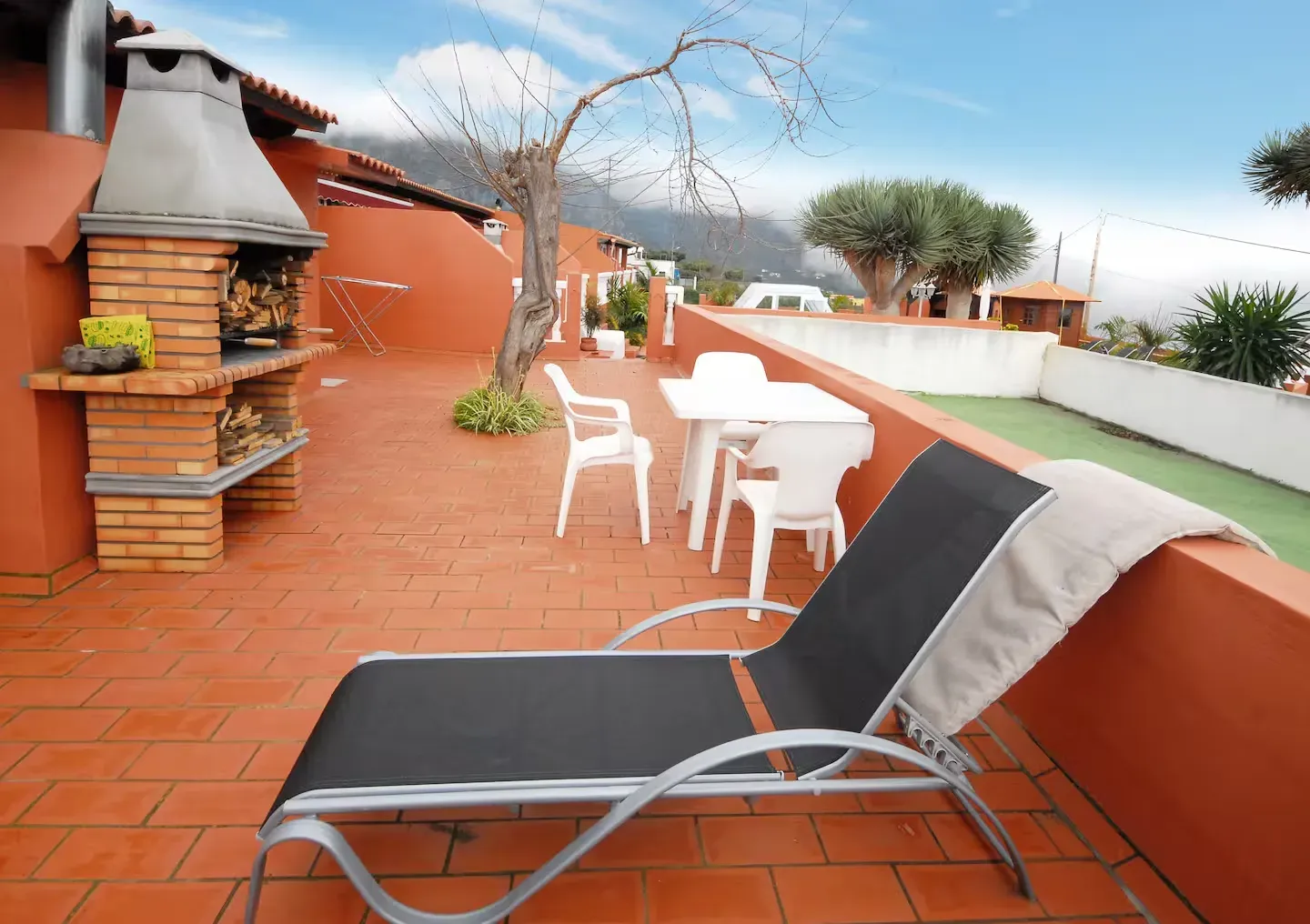Un patio exterior con suelo de baldosas de terracota, que cuenta con una barbacoa de ladrillo, una tumbona con una toalla blanca y un juego de comedor.