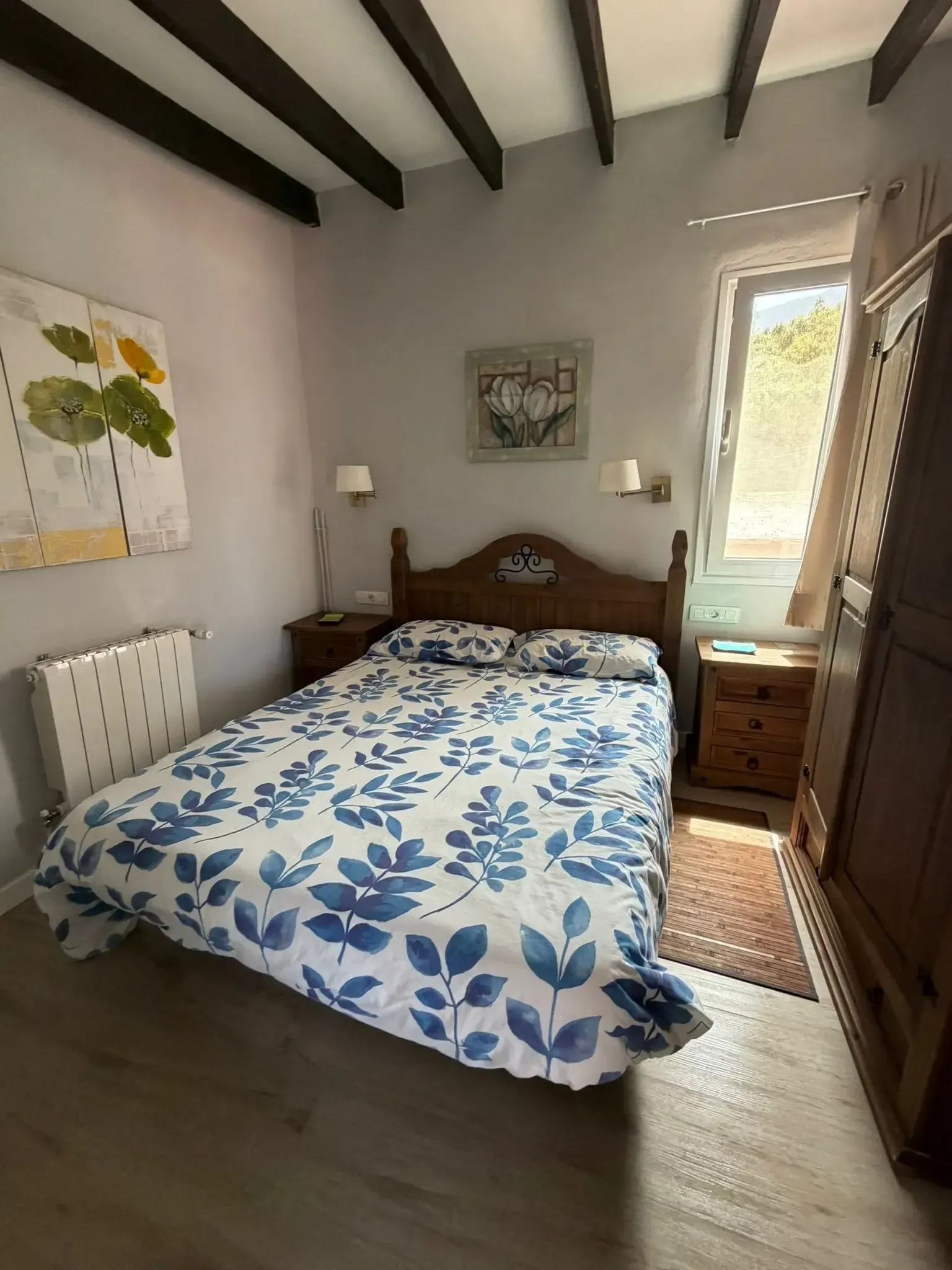 Un dormitorio con cama doble, edredón con estampado de hojas blancas y azules, muebles de madera y vigas de techo a la vista.