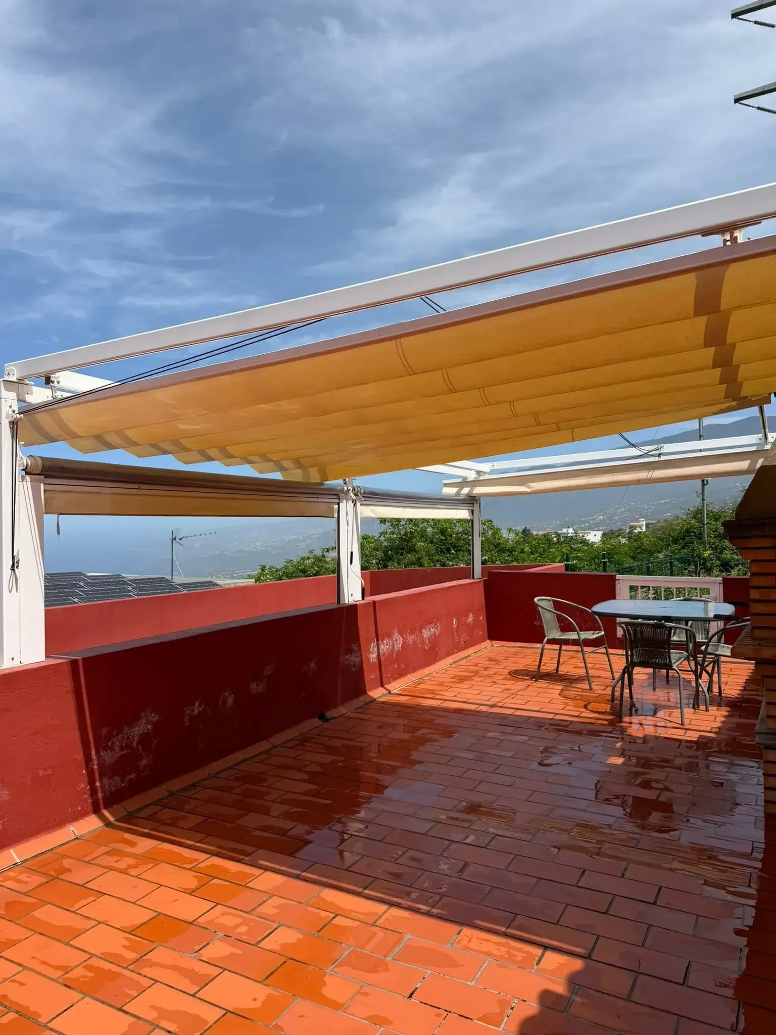 Una pérgola retráctil de color beige da sombra a un patio húmedo con baldosas de terracota y asientos al aire libre, con las colinas lejanas como telón de fondo.