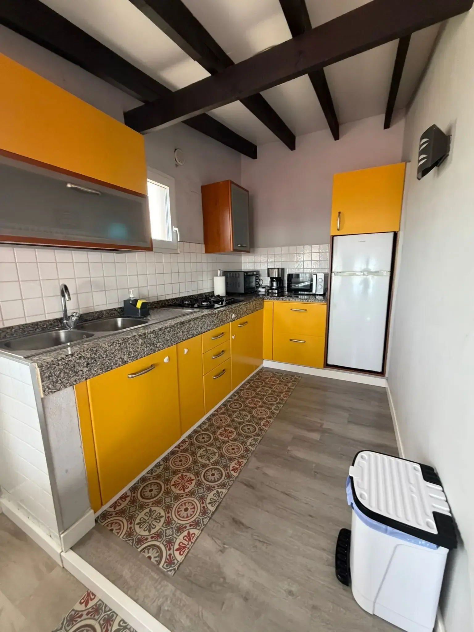 Una cocina estrecha y moderna con armarios de color amarillo vibrante, encimeras grises, azulejos blancos tipo metro en la pared y vigas de madera oscura.