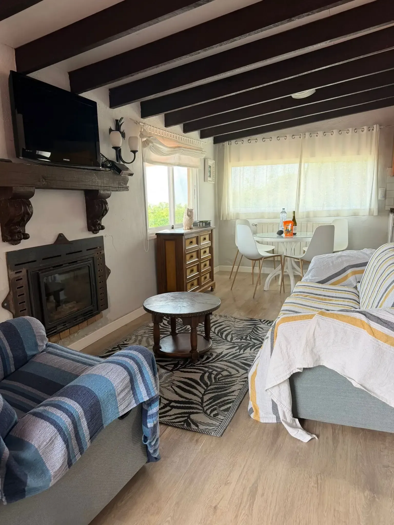 Una acogedora sala de estar con chimenea, vigas de madera oscura en el techo, muebles con estampados azules y un comedor junto a la ventana.