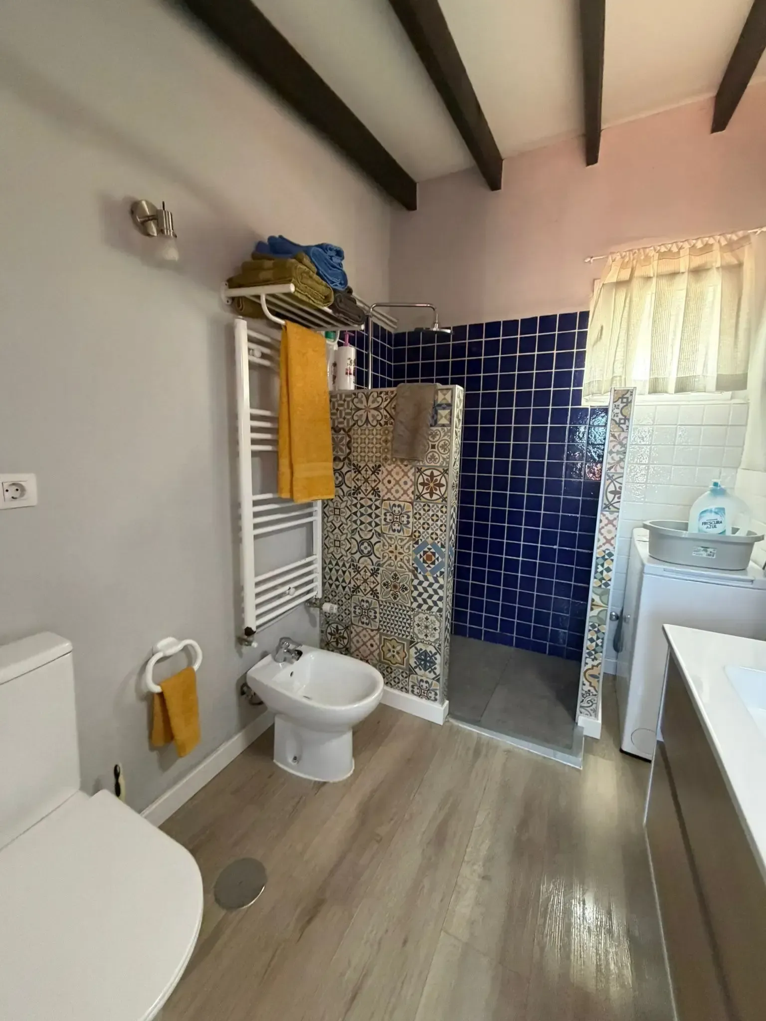 Un baño moderno con bidé, inodoro, toallero con toallas amarillas, pared de ducha con azulejos estampados y suelo de madera.