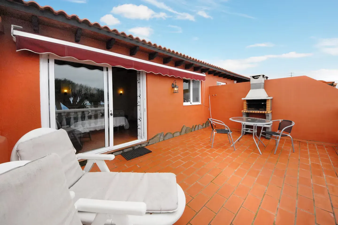 Un patio con baldosas de terracota, muebles de salón blancos, un pequeño juego de comedor y una parrilla exterior contra una pared naranja.