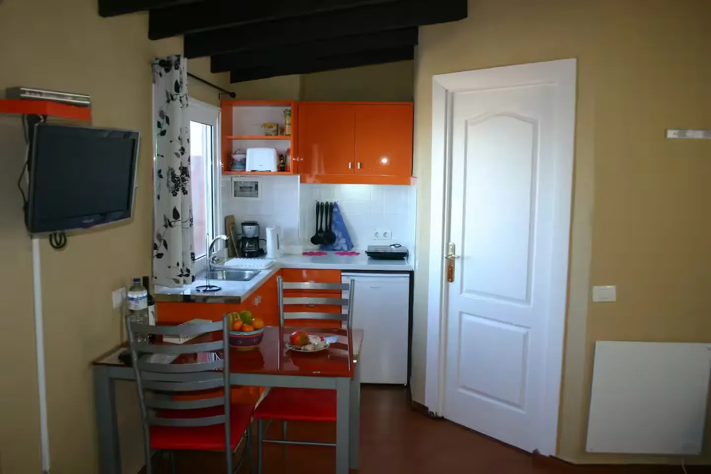 Una pequeña cocina con armarios de color naranja brillante, una mesa de comedor con dos sillas rojas, un televisor montado en la pared y una puerta blanca.
