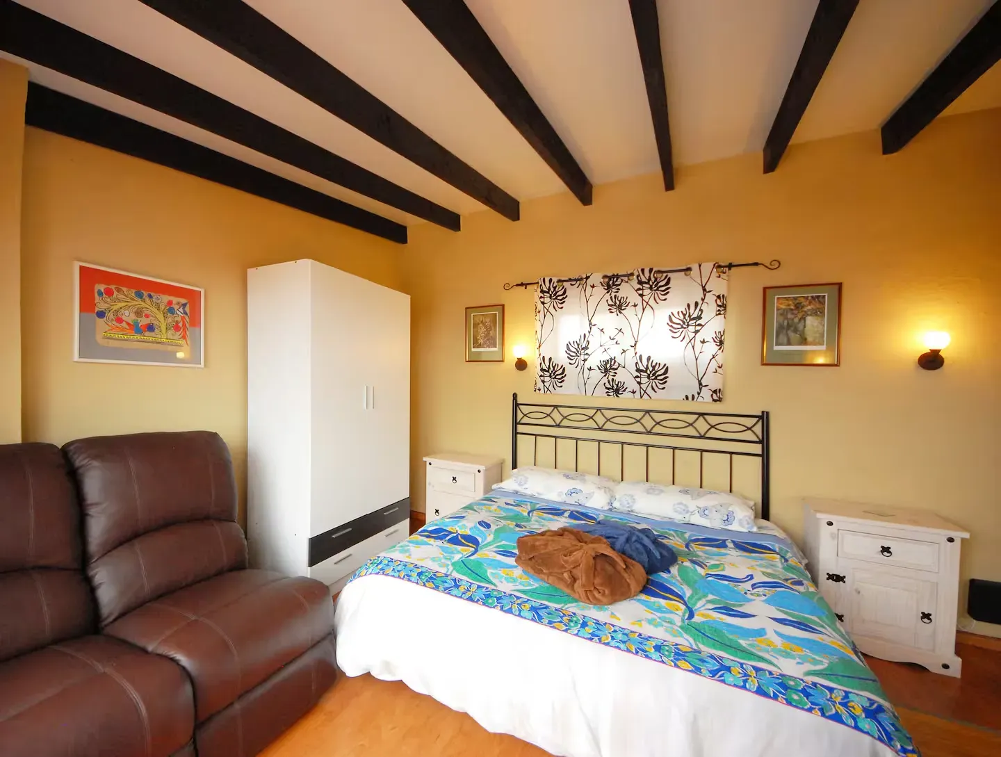 Un dormitorio con paredes amarillas, vigas oscuras en el techo, una cama con una colcha azul estampada, un sillón marrón y muebles blancos.