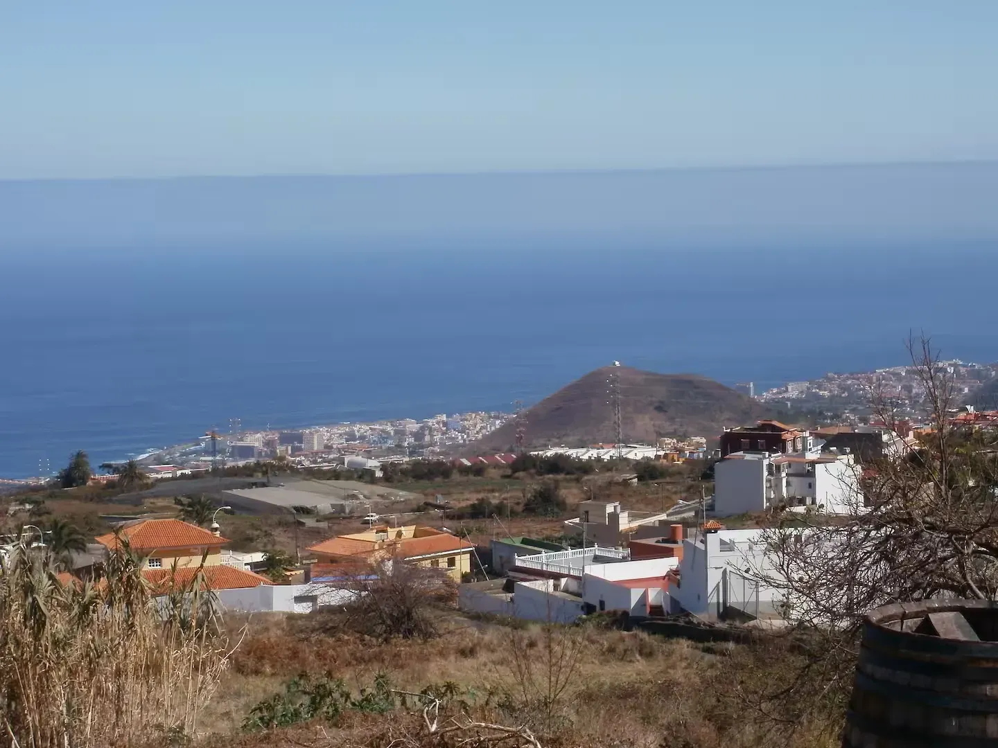Una vista panorámica de un pueblo costero al pie de una colina volcánica, con vistas al océano azul bajo un cielo despejado.