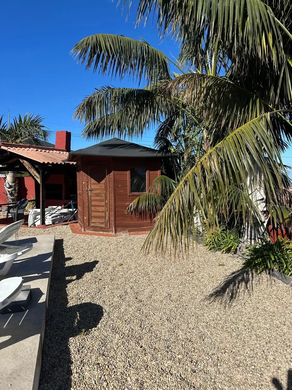 Un cenador de madera marrón se alza en un patio de grava junto a una terraza cubierta, bajo un cielo azul brillante, con una palmera.