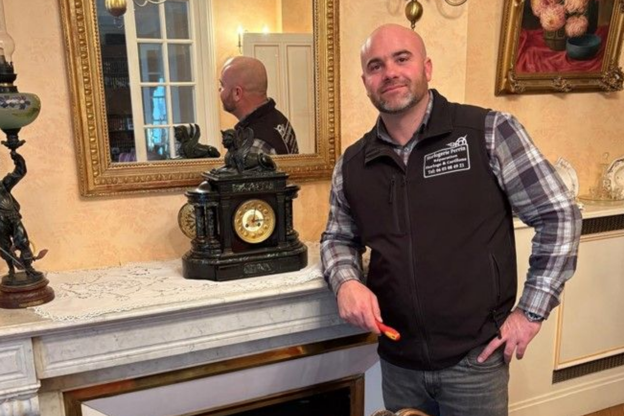 Un homme se tient près d'une cheminée où est posée une horloge. Miroir doré et décor orné en arrière-plan.