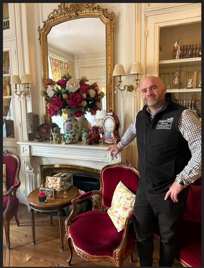Un homme en gilet pose près d'un fauteuil orné et d'une cheminée.