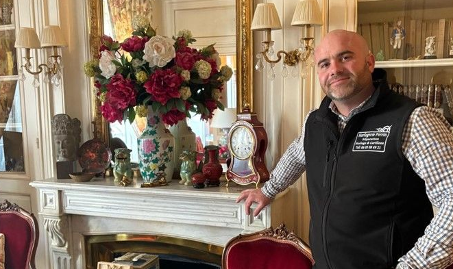 Un homme en gilet se tient près d'une cheminée ornée de fleurs et d'une horloge.