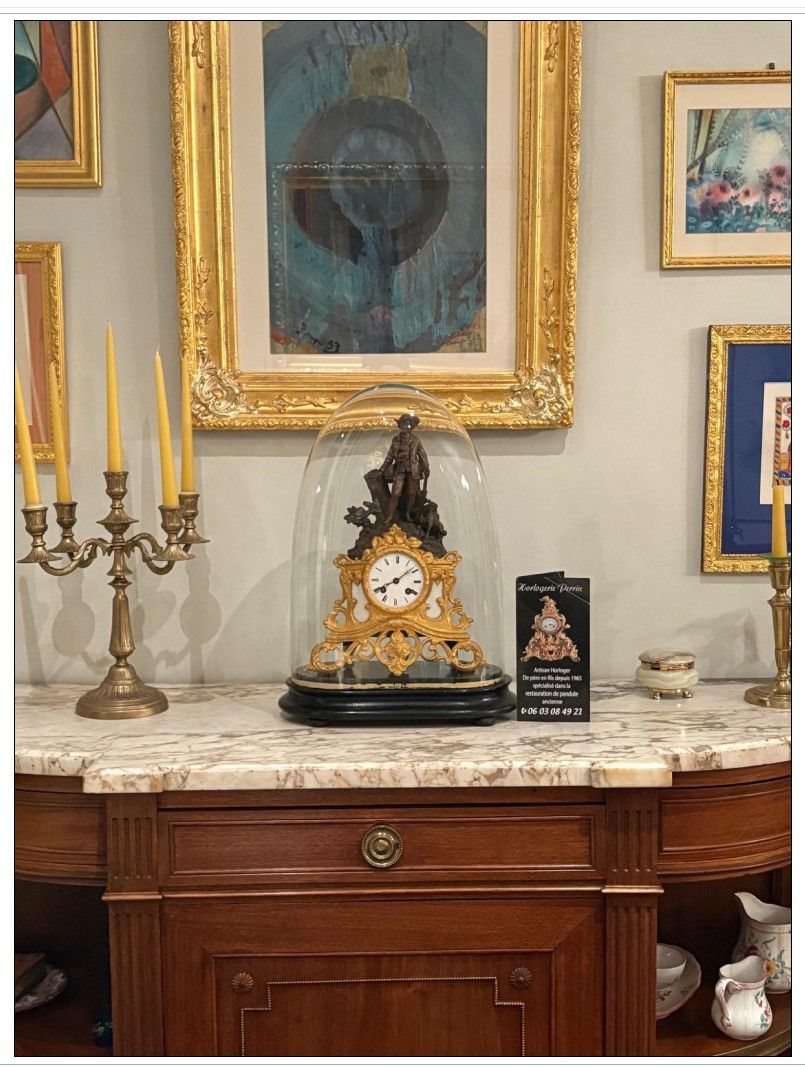 Une horloge ancienne sous une cloche de verre, posée sur un meuble en bois.