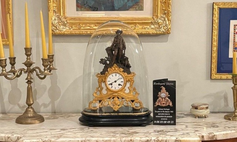 Horloge sous cloche de verre sur marbre. Bougeoir à gauche, tableau encadré en arrière-plan.