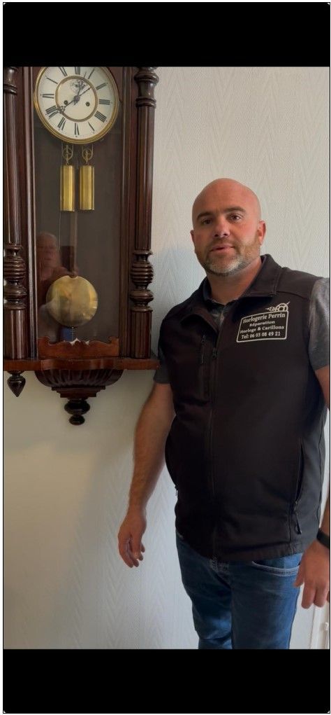 Un homme se tient à côté d'une horloge murale en bois. Il porte un gilet noir, un jean et une montre.