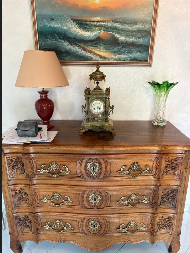 Commode en bois ornée de sculptures décoratives, surmontée d'une lampe, d'une horloge et d'un vase.