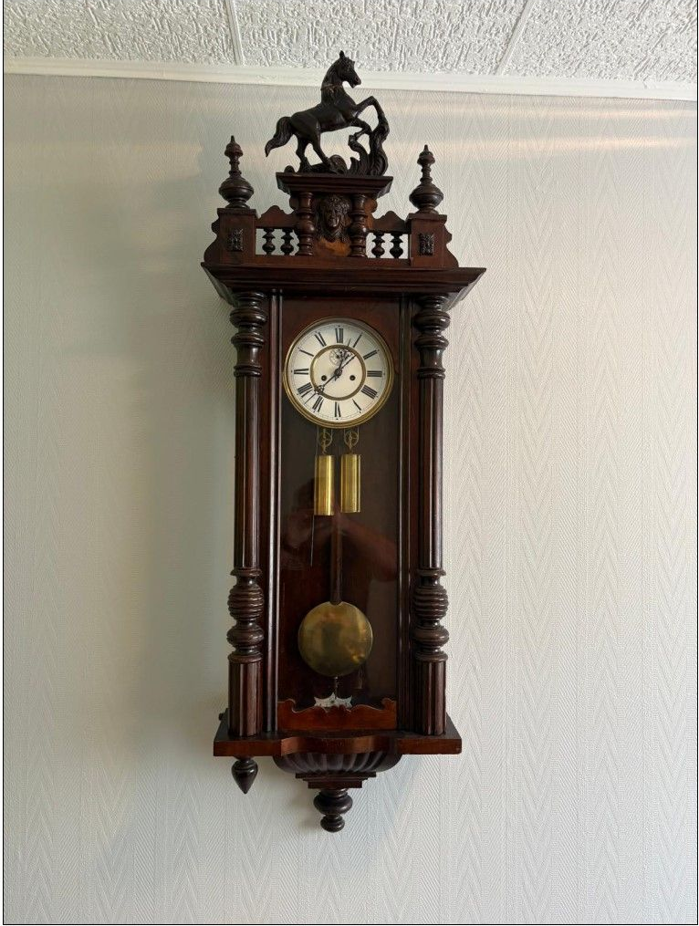 Horloge murale en bois surmontée d'un cheval, sur un mur gris clair.