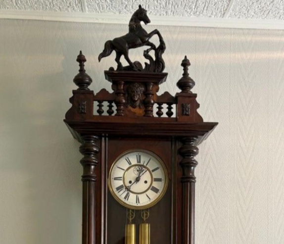 Horloge ancienne en bois surmontée d'une sculpture de cheval. Cadran blanc, pendule visible.