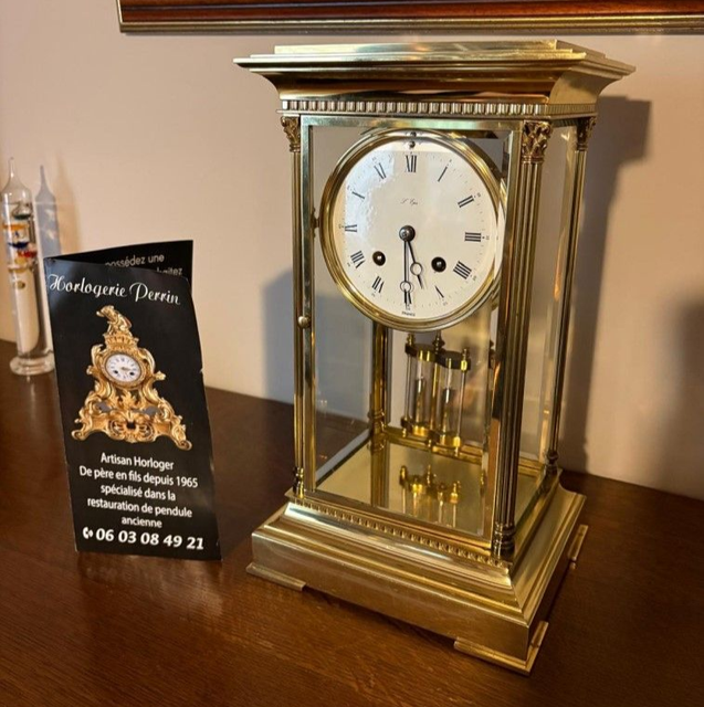 Horloge dorée à cadran blanc et chiffres romains, sous verre. Posée sur une table à côté d'une carte de visite.