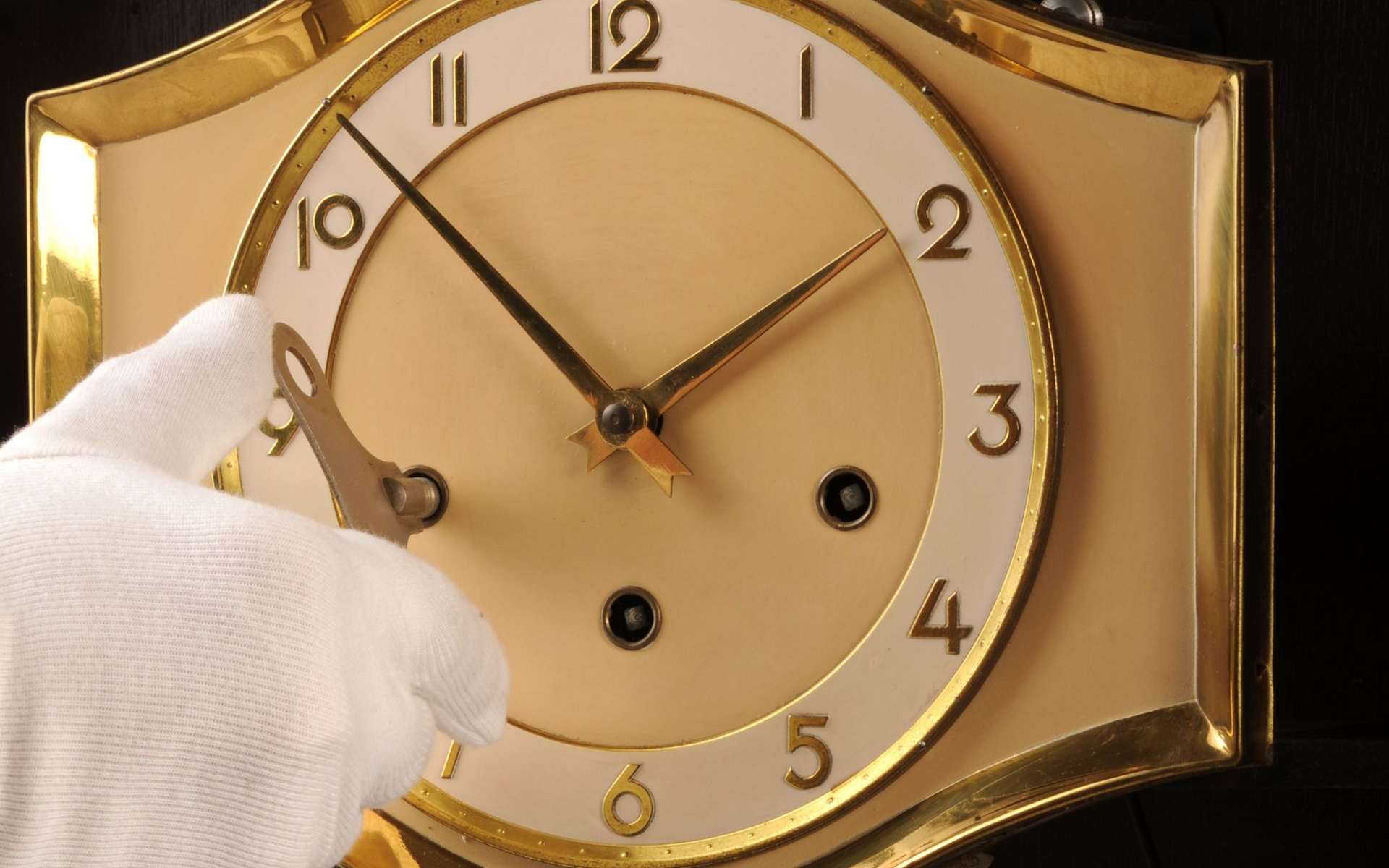 Main avec un gant blanc remontant une horloge à cadre doré avec une clé ; le cadran de l'horloge indique 10 h 10.