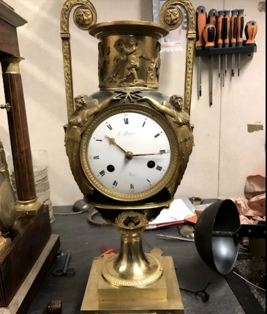 Pendule ancienne en or en forme de vase, personnages ornés, cadran blanc avec aiguilles et pièces manquantes.