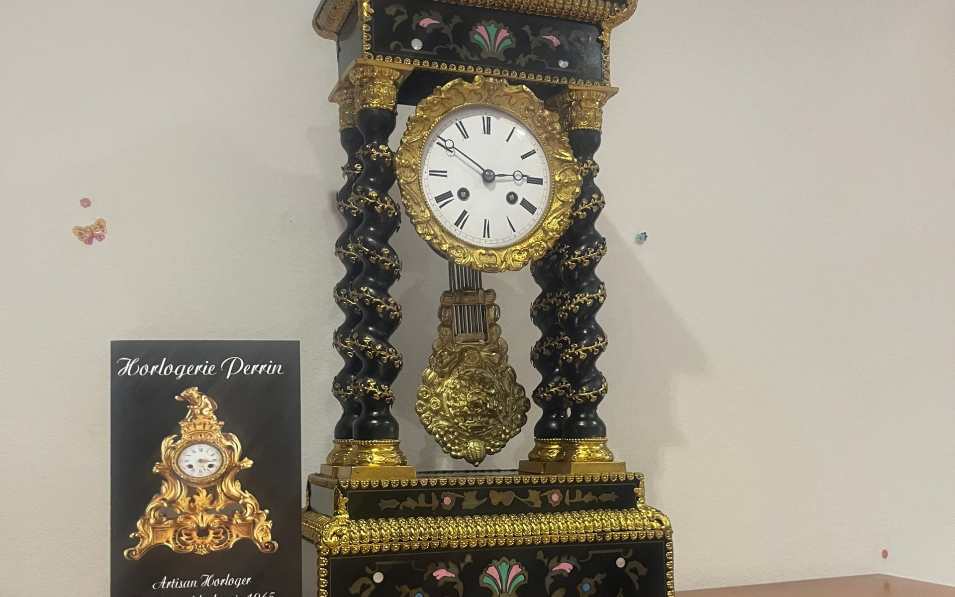 Horloge ancienne avec garniture dorée, base noire et détails ornés, avec une image plus petite sur la gauche.