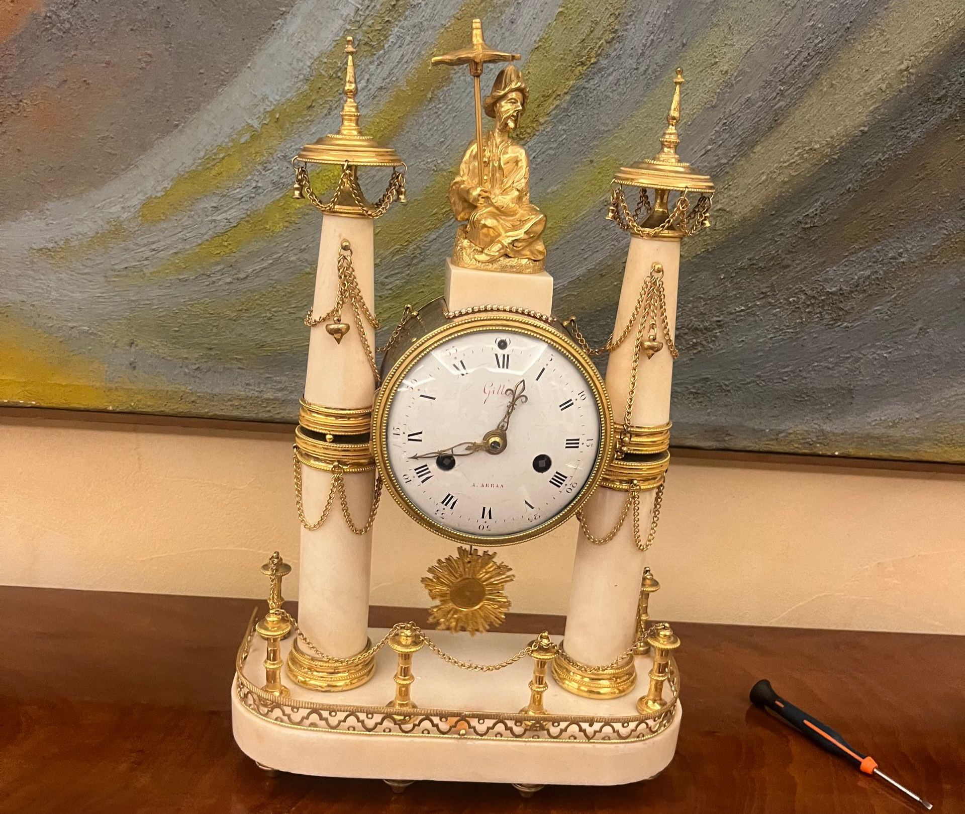 Horloge ancienne avec des accents dorés, des colonnes blanches, un personnage en haut et un tournevis sur le côté.