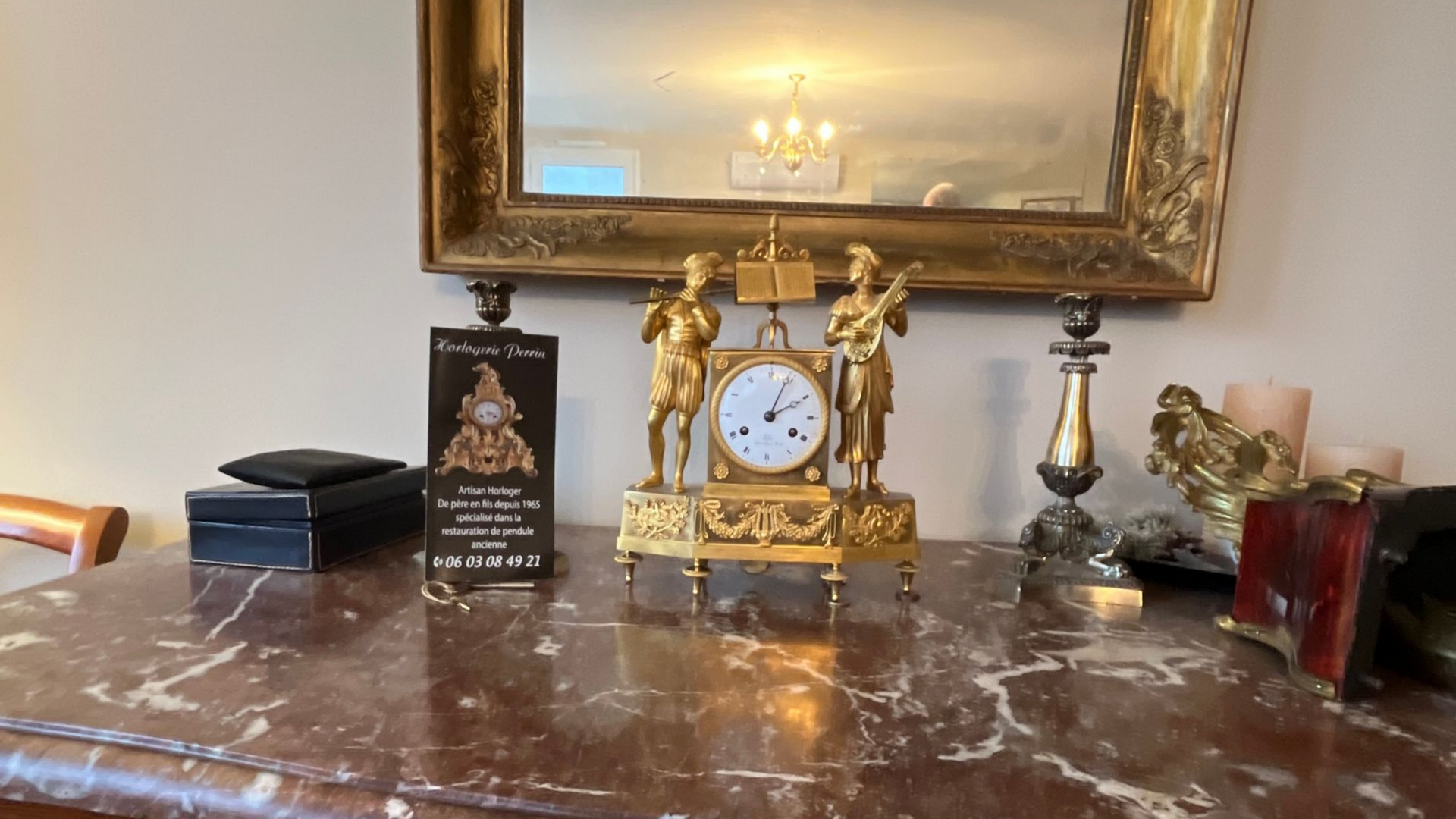 Horloge ancienne en or avec deux personnages sur une table en marbre, sous un miroir encadré d'or.