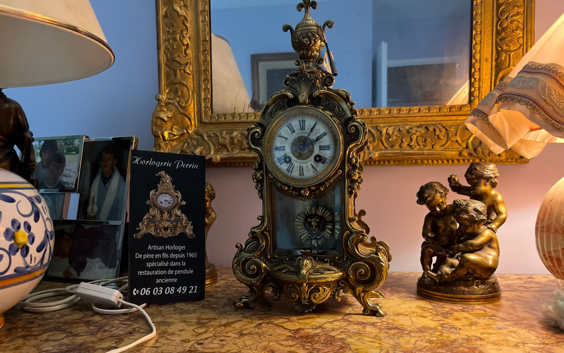 Horloge antique dorée sur une surface en marbre, flanquée de lampes, de statues et de photos encadrées.