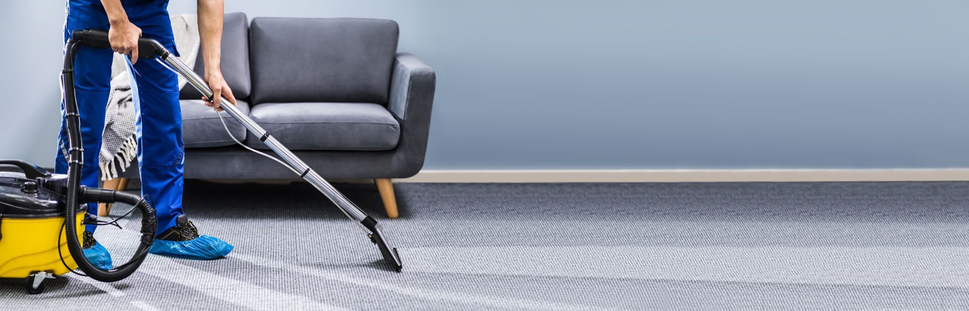 Una persona che utilizza un pulitore per tappeti su un pavimento in moquette vicino a un divano grigio.