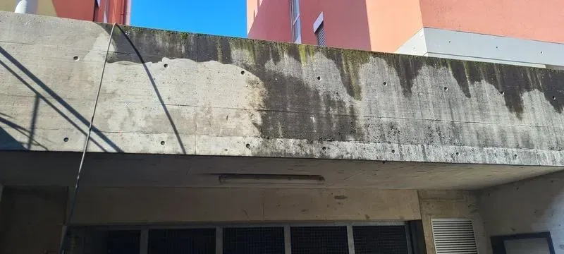 Struttura in cemento con macchie d'acqua, sotto un cielo azzurro, ed edifici adiacenti.