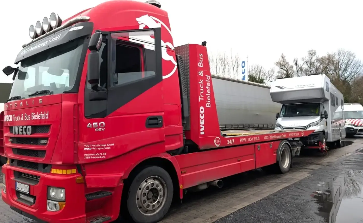 Ein roter Autotransporter verlädt ein weißes Fahrzeug auf der Straße; dahinter fährt ein weiterer Lkw.