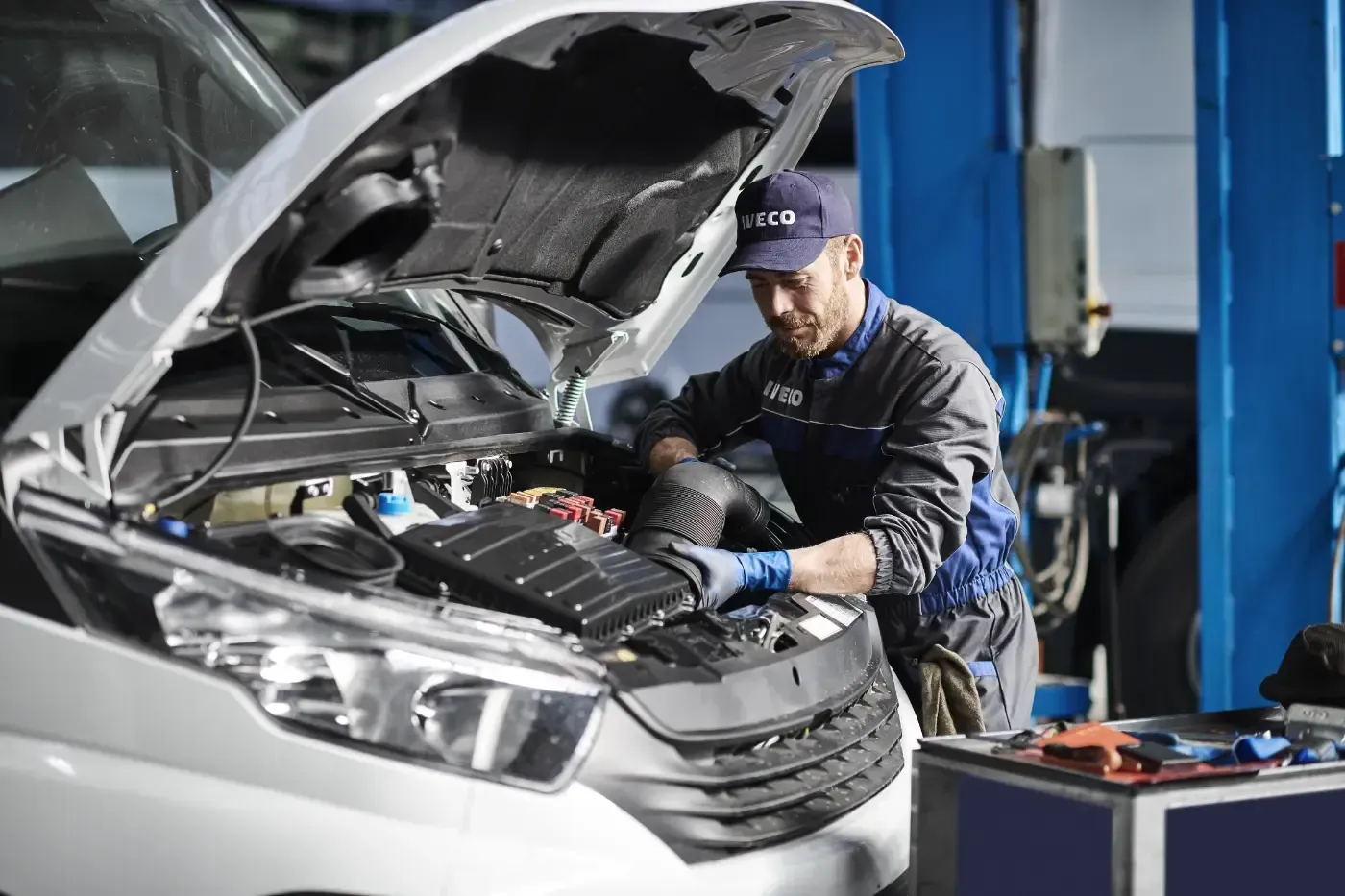 Ein Mechaniker arbeitet in einer Werkstatt unter der geöffneten Motorhaube eines weißen Autos.