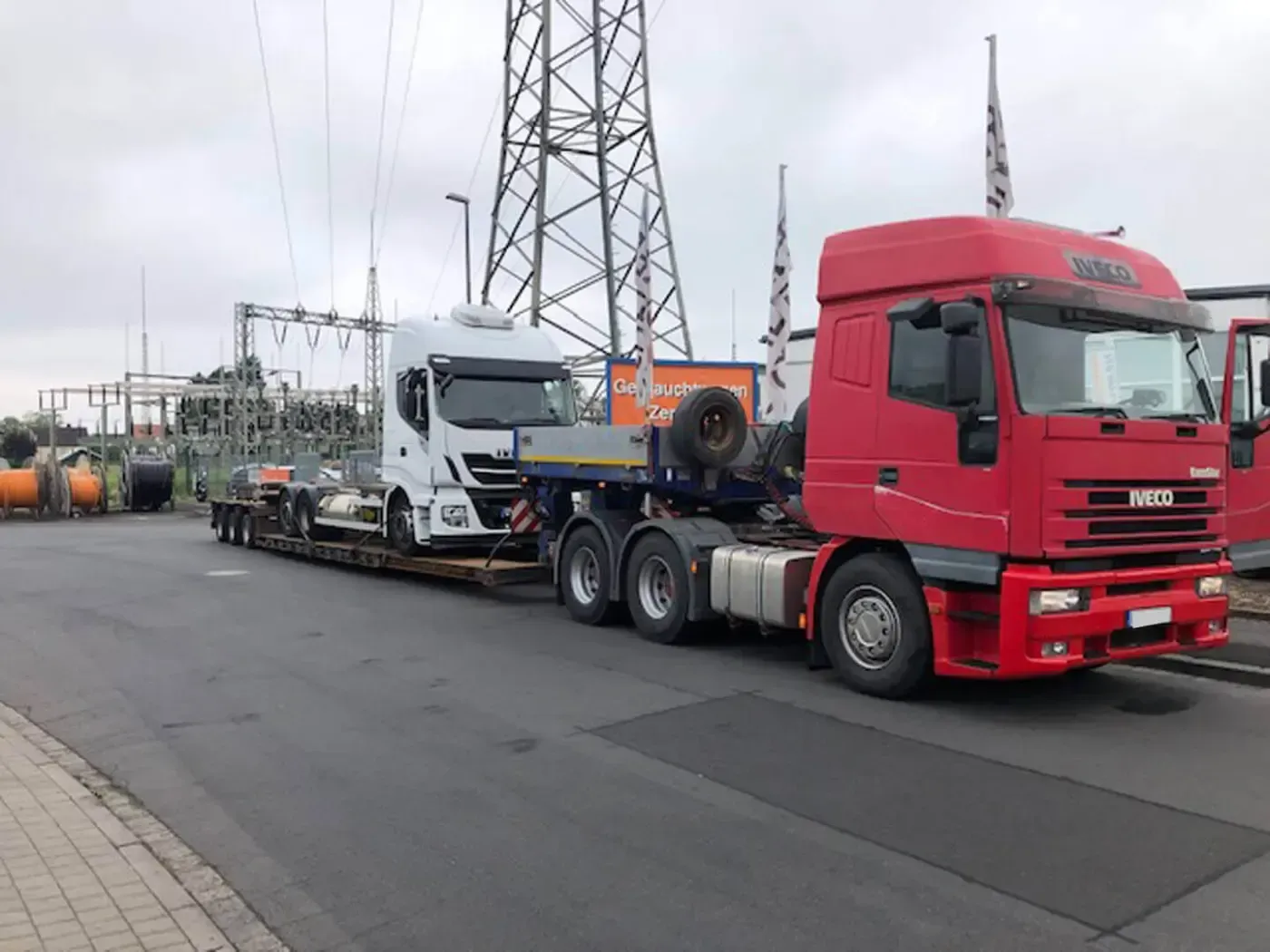 Ein roter Lastwagen mit einem weißen Lastwagen als Anhänger steht auf einem Industriegelände.