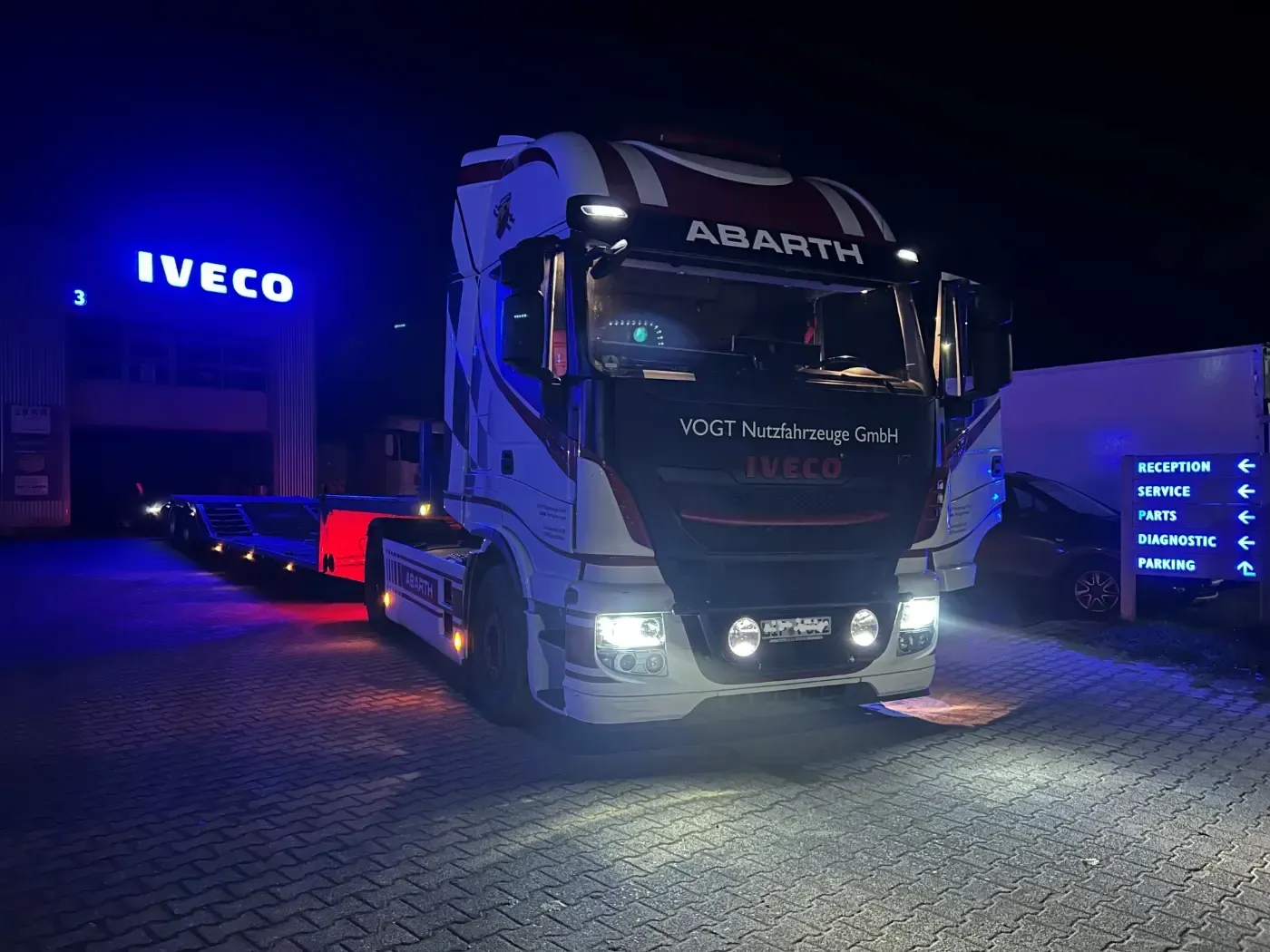 Ein blauer Iveco Abarth-Lkw bei Nacht, geparkt auf Kopfsteinpflaster, mit beleuchteter Markenbeschriftung und eingeschalteten Scheinwerfern.