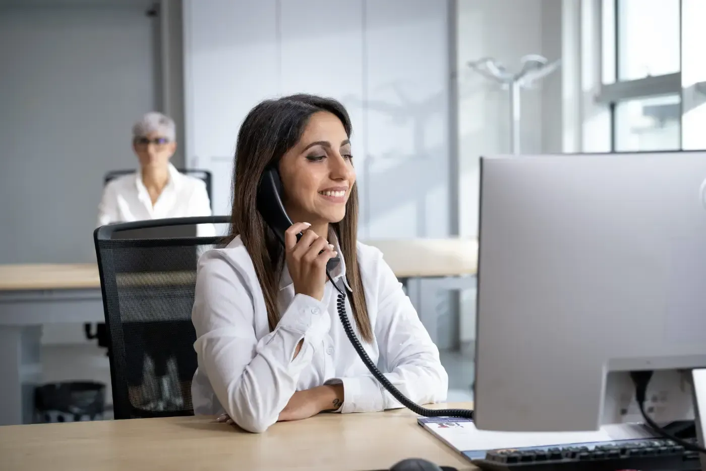 Eine Frau telefoniert an einem Tischtelefon an einem Büroarbeitsplatz; im Vordergrund ist ein Computermonitor zu sehen.