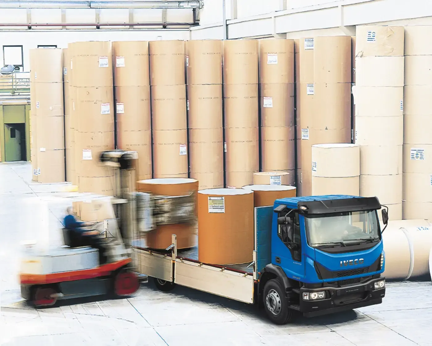 Ein blauer Lastwagen transportiert große Kartonrollen in einem Lagerhaus, in der Nähe fährt ein Gabelstapler.