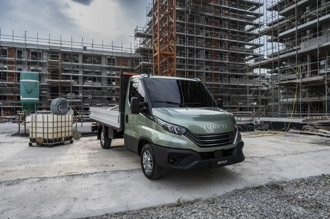 Ein grüner Lieferwagen parkt auf einer Baustelle; im Hintergrund sind Gerüste und Betongebäude zu sehen.