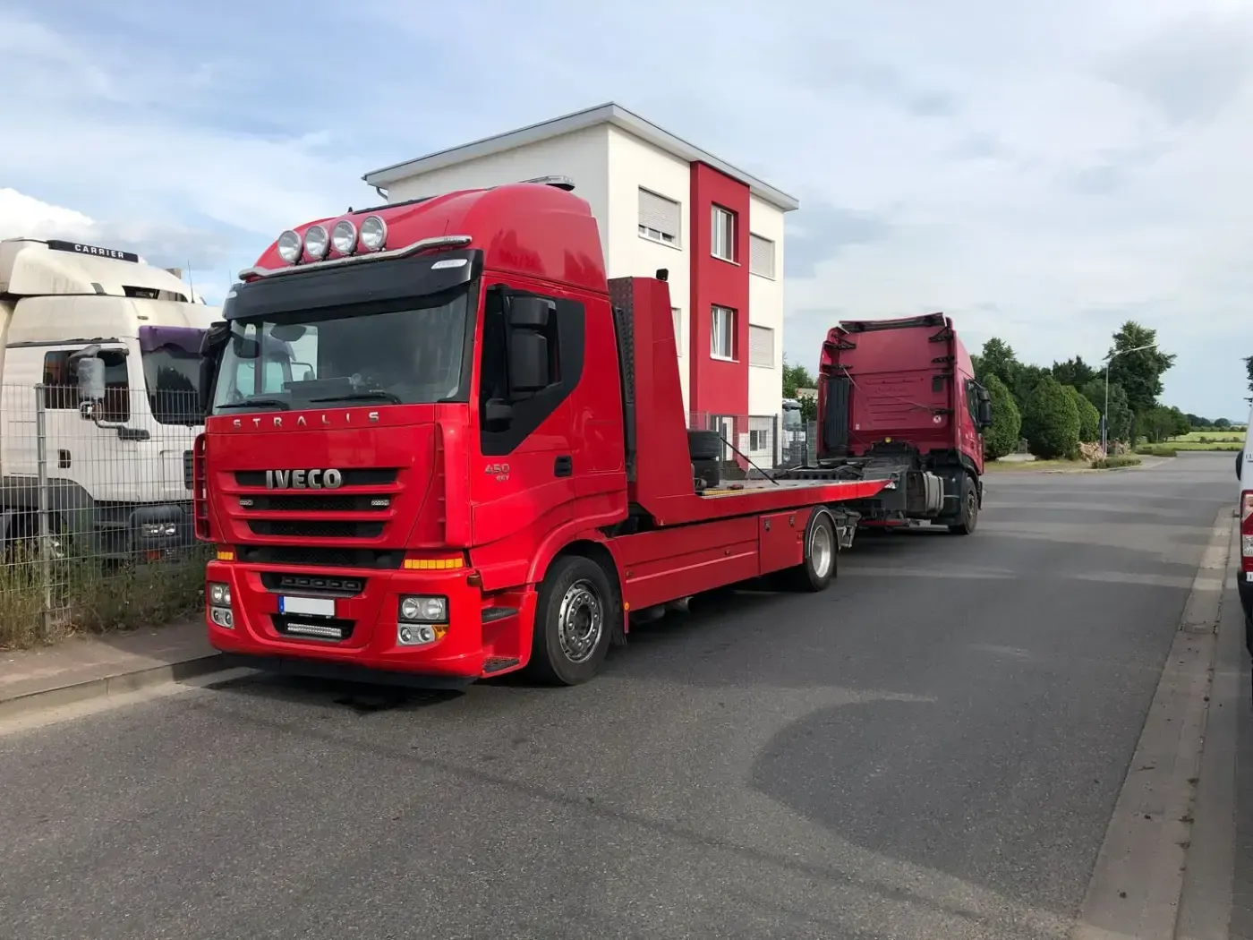 Ein roter Abschleppwagen mit Pritsche parkt am Straßenrand, dahinter ein weiterer Lkw unter bewölktem Himmel