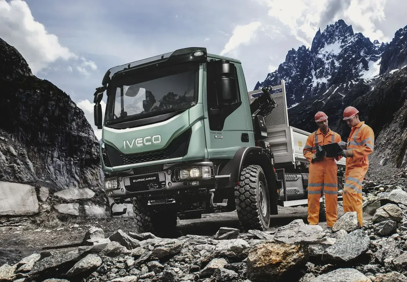 Zwei Arbeiter in orangefarbener Schutzkleidung neben einem grünen Iveco-Lkw auf einer felsigen Bergstraße mit schneebedeckten Gipfeln.