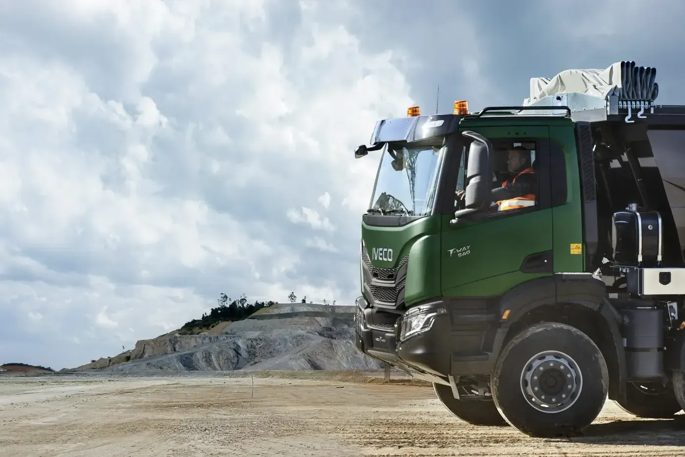Grüner Muldenkipper auf einem Feldweg in der Nähe einer felsigen Baustelle unter bewölktem Himmel