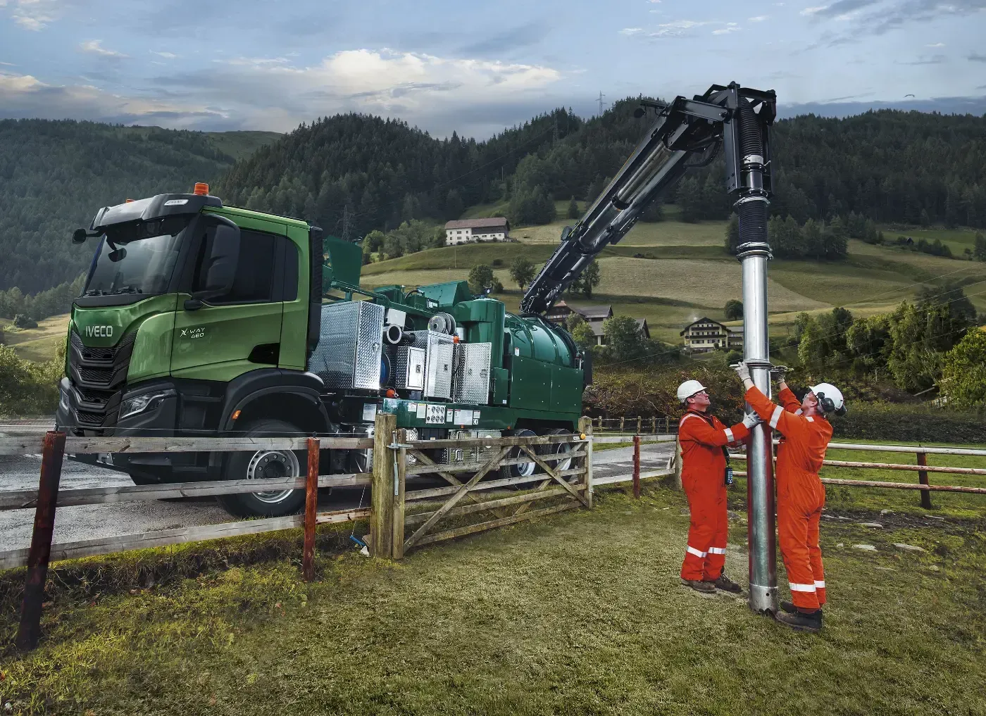 Arbeiter in orangefarbenen Schutzanzügen inspizieren einen grünen, auf einem LKW montierten Kran am Straßenrand einer ländlichen Straße.