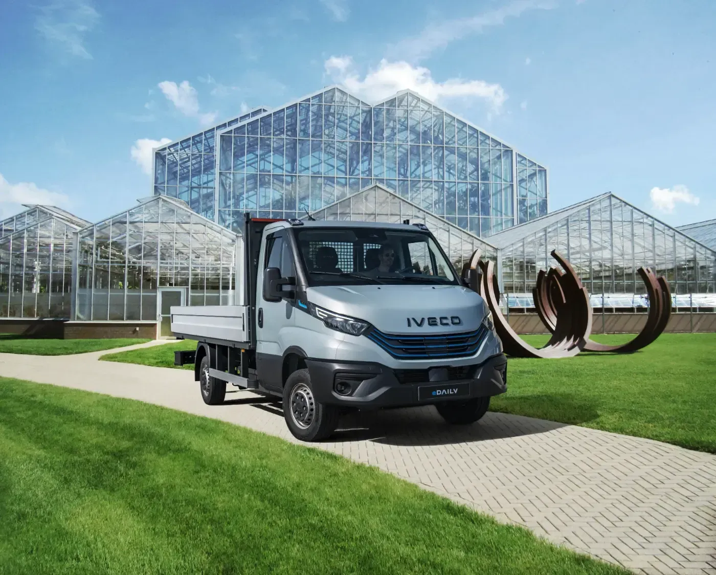 Ein silberner IVECO-LKW parkte auf einem Weg vor großen Glasgewächshäusern auf einem sonnigen Rasen.