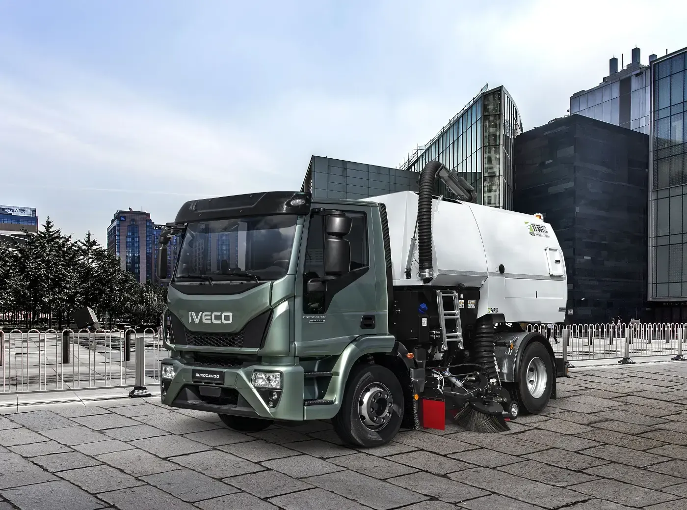 Ein grüner und weißer Iveco-Müllwagen parkt auf einem Stadtplatz; im Hintergrund sind moderne Gebäude zu sehen.