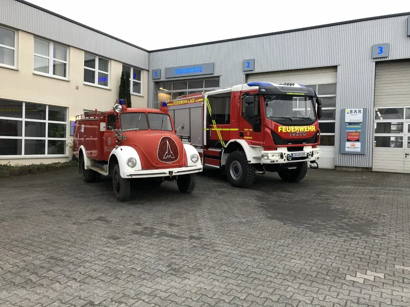 Zwei rote Feuerwehrwagen parkten vor einer Feuerwache auf einem nassen Kopfsteinpflasterplatz.