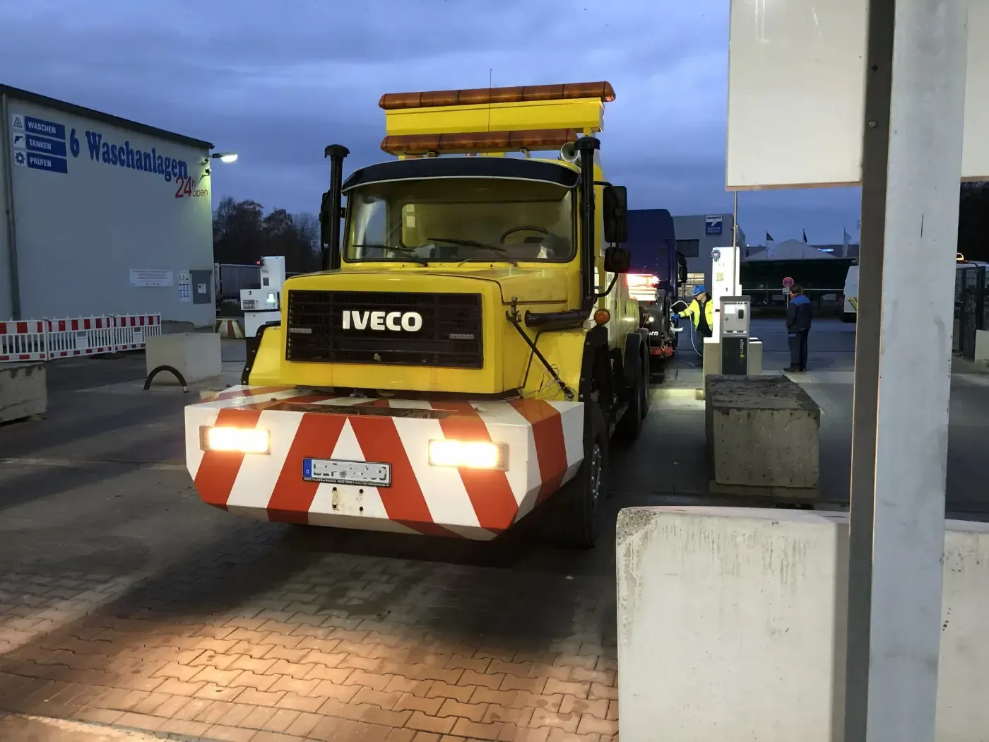 Ein gelber Iveco-Nutzwagen mit rot-weiß gestreifter Stoßstange parkt in der Abenddämmerung an einer Tankstelle.