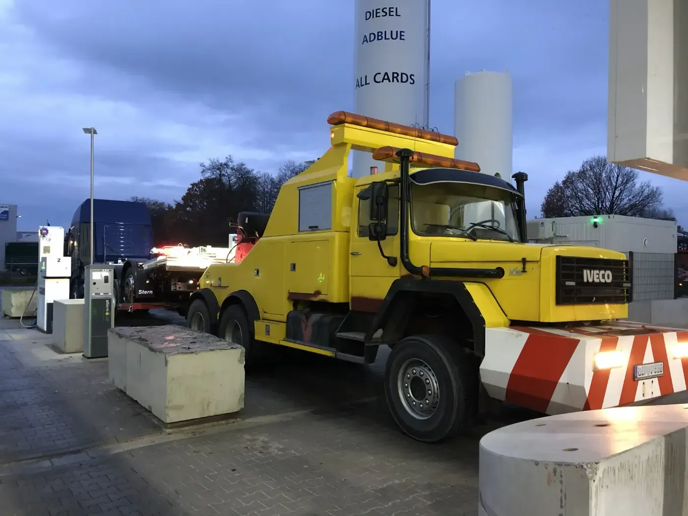 Ein gelber Abschleppwagen parkt in der Abenddämmerung an einer Zapfsäule einer Tankstelle; davor befindet sich eine orange-weiße Absperrung.