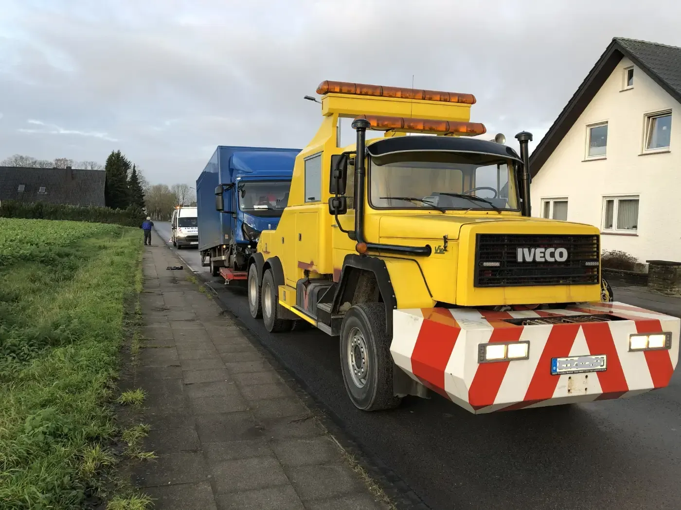 Ein gelber Abschleppwagen zieht einen blauen Lkw mit rot-weiß gestreifter Frontstoßstange in einer Wohnstraße ab.