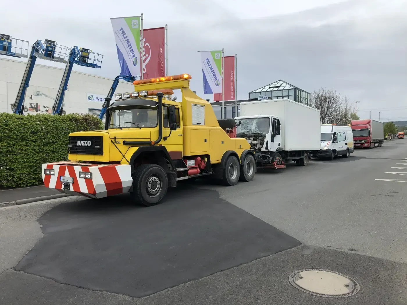 Ein gelber Abschleppwagen zieht einen weißen Kastenwagen durch eine Stadtstraße mit Flaggen und Gebäuden in der Nähe.