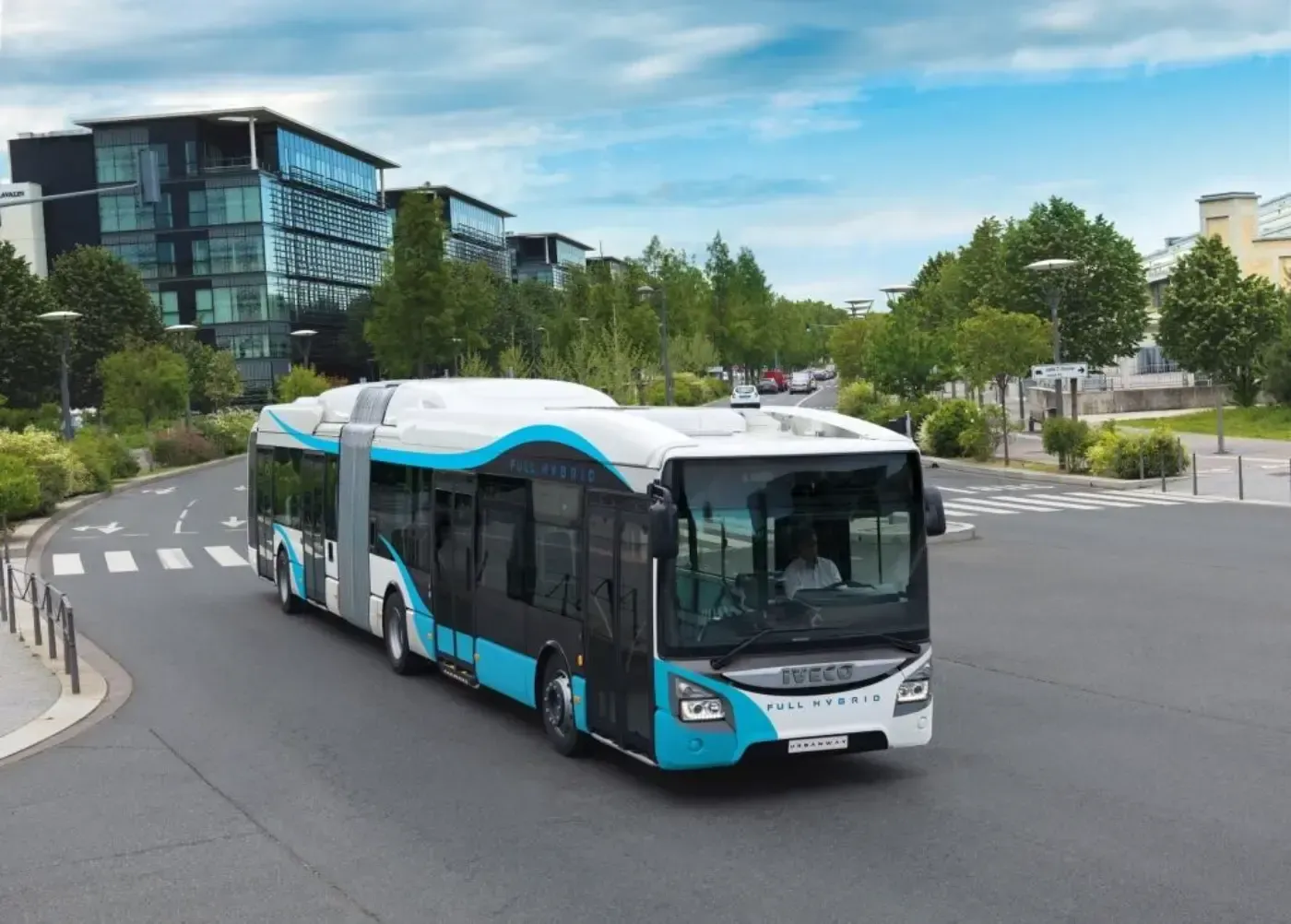 Ein blau-weißer Stadtbus biegt in einer kurvigen Straße in der Nähe moderner Gebäude und Bäume ab.