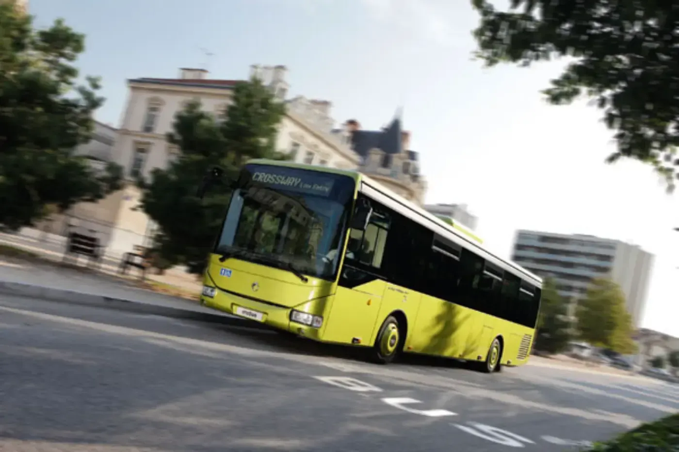 Ein gelber Stadtbus biegt in eine Straße ein, im Hintergrund sind Gebäude und Bäume zu sehen.